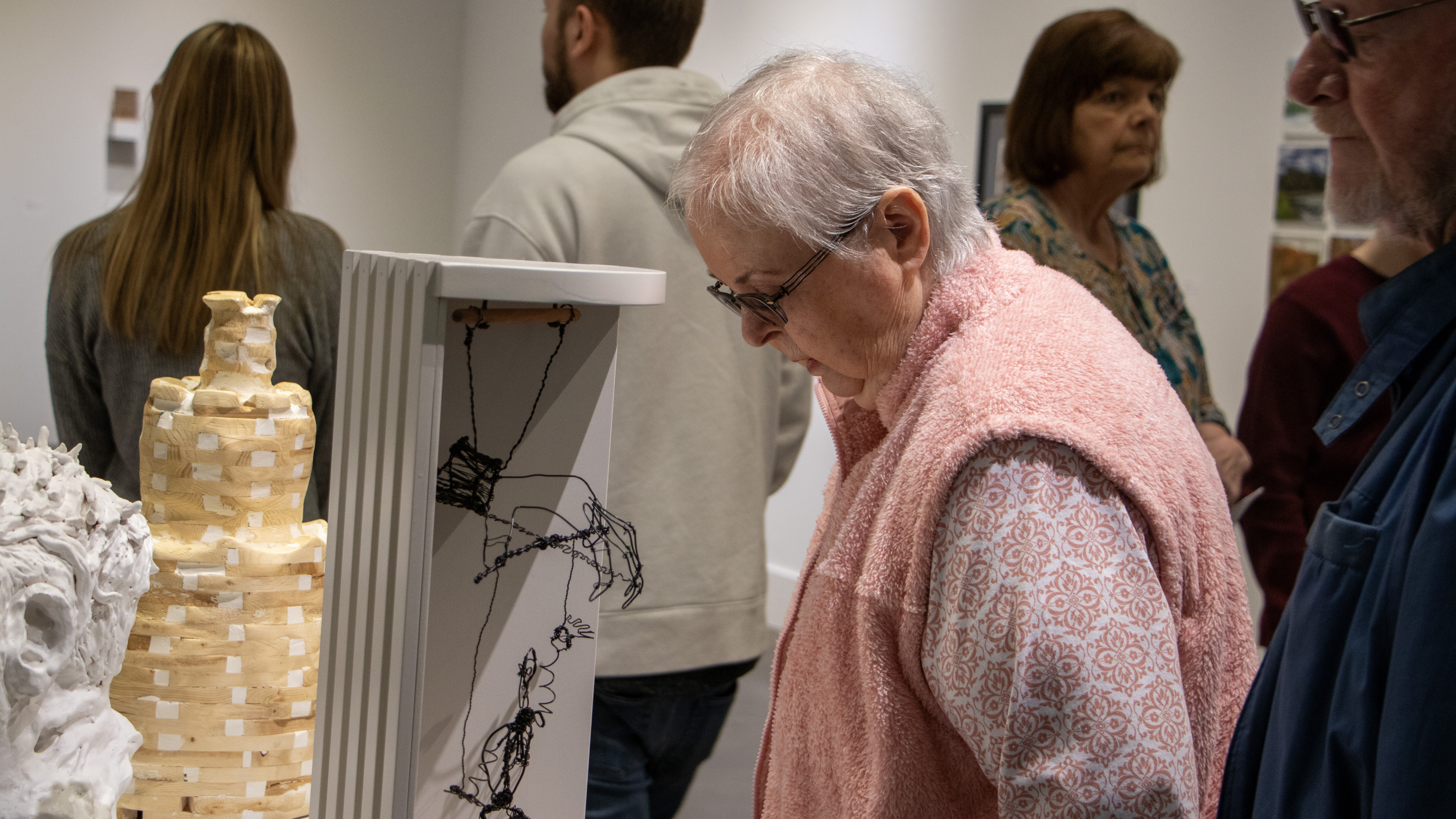 an older woman bends slightly to look down at a wire and sculptural work